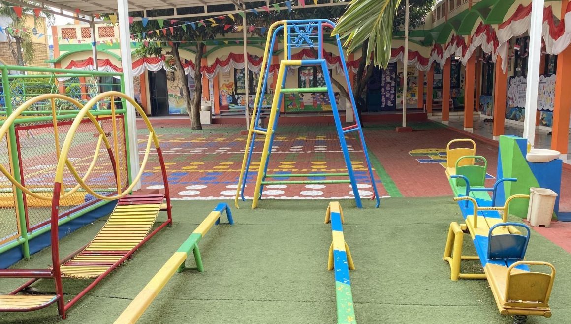 Taman bermain beralas rumput sintetis dengan tangga panjat, titian, dan jungkat-jungkit