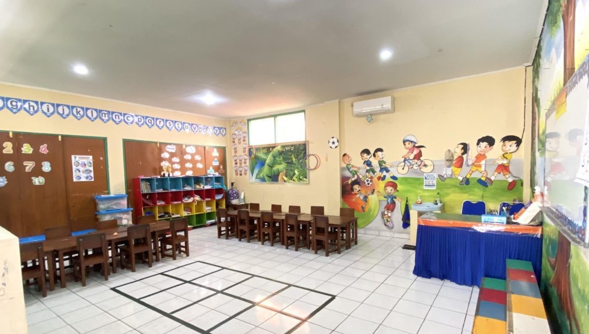 Ruang kelas dengan meja dan kursi kayu, rak mainan, dan mural anak bermain