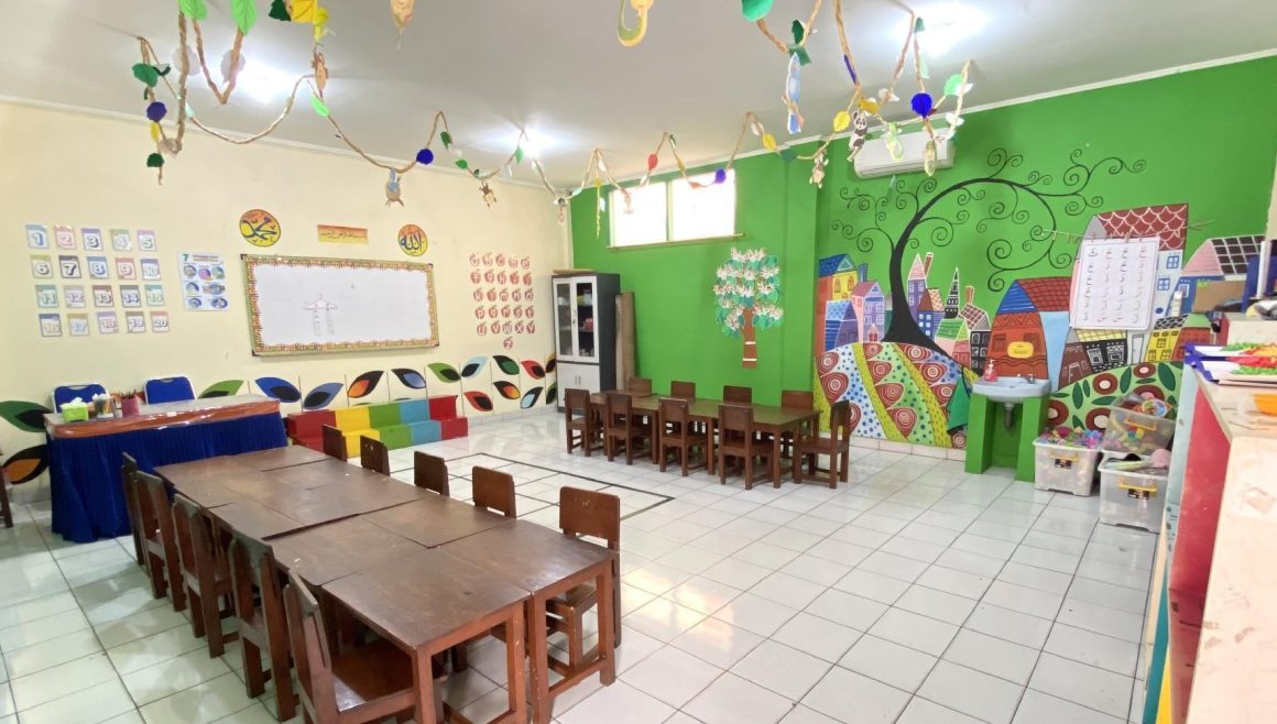 Ruang kelas berdinding hijau bermural pohon dengan meja kayu panjang